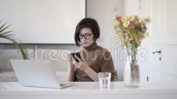 亚洲女人坐在白色的家庭办公室里滚动电话视频的预览图