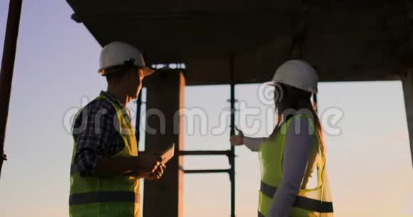 建筑工人拿着一块石碑一个戴着白盔的女检查员站在屋顶上在日落时分握手视频的预览图