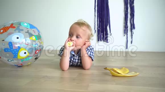 粗心的蹒跚学步的男孩躺在地板上吃黄色的香蕉万向节运动视频的预览图