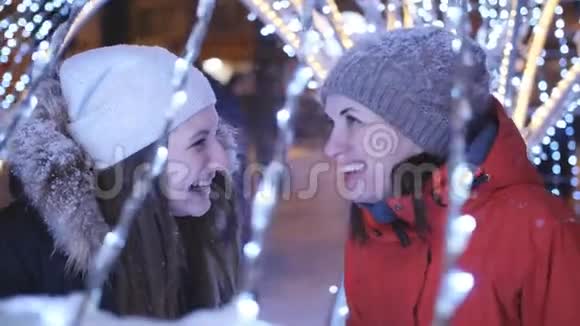 两个女孩在一个冬天的晚上漫步在市中心为圣诞节装饰了明亮的灯光和花环视频的预览图