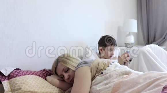 疲惫的母亲在睡梦中醒来太阳坐在床上用小工具智能手机玩手机游戏视频的预览图