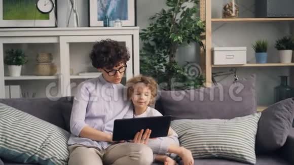 母亲和儿子在舒适的房子里用平板电脑看屏幕在沙发上说话视频的预览图