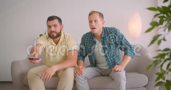 两位成年白种人男性朋友在电视上看足球时被迷倒坐在电视上的特写镜头视频的预览图