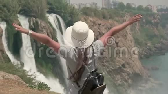 一个戴着帽子和背包的旅游女孩站在瀑布前双臂张开视频的预览图
