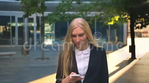 开朗的商务女性在户外打电话穿着黑色夹克的金发女人拿着智能手机看着远方视频的预览图