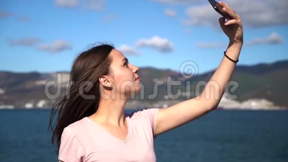 迷人的年轻女性使用手机在海滩上自拍背景是山脉和多云的天空视频的预览图
