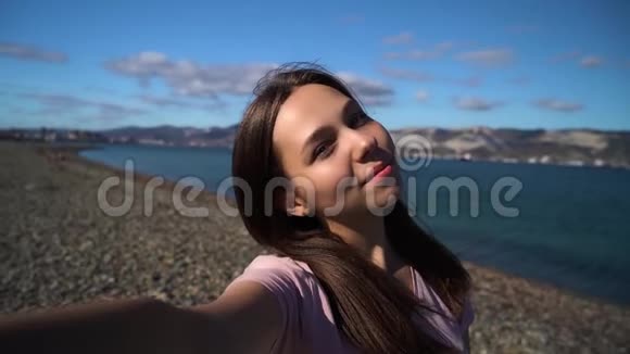 迷人的年轻女性使用手机在海滩上自拍背景是山脉和多云的天空视频的预览图