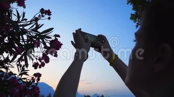 一个女孩在日落时在智能手机上拍摄花的照片背景是山视频的预览图