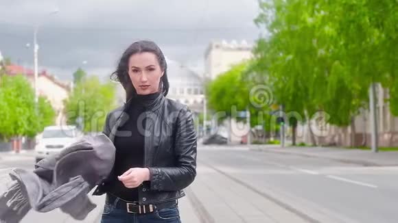 年轻漂亮的女孩穿着黑色皮夹克在城市的背景下拿着一条灰色的围巾在胜利中飘扬的头发视频的预览图