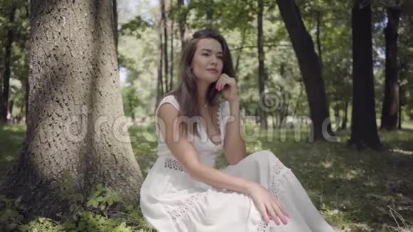 迷人的年轻女孩的肖像长着一头深色长发穿着一件白色的夏装坐在树下视频的预览图