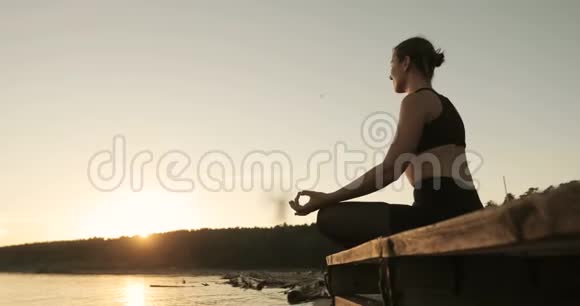 年轻的牧人正坐在河边的码头上坐在夕阳下的莲花姿态视频的预览图