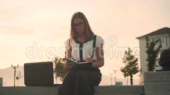 商务女性穿着白色衬衫和眼镜坐在长凳上工作女孩写在笔记本里看着笔记本电脑视频的预览图
