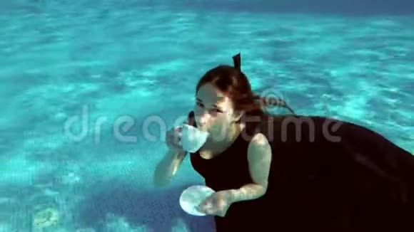 可爱的女孩穿着裙子坐在池底水下她从白色的杯子里喝下酒浮到水面上视频的预览图