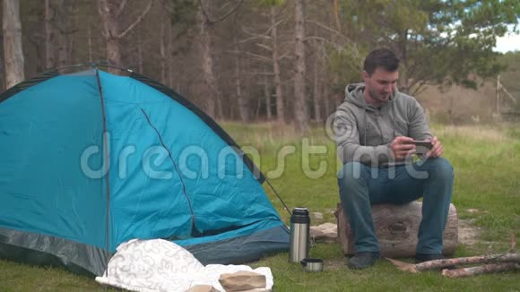 一个年轻人坐在蓝色帐篷附近的一根大木头上在电话上拍了一张照片视频的预览图