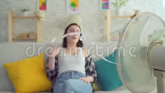 年轻女子坐在沙发上手里拿着电风扇从暖气里逃出来视频的预览图