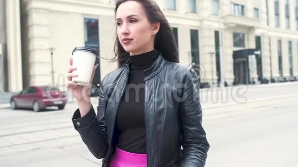 美丽的年轻女孩穿着黑色皮夹克在一条城市街道的背景下咬着太阳镜饮料咖啡视频的预览图