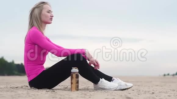 美丽的年轻运动女子在海滩上训练后休息从塑料瓶里喝水视频的预览图