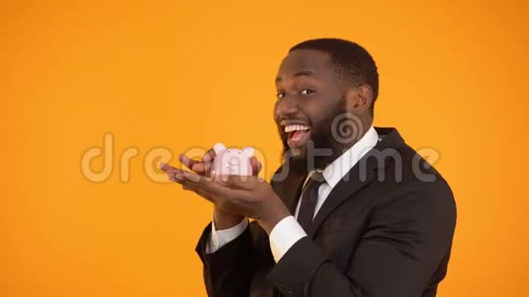 快乐的黑人男性穿着正式服装摇着猪储蓄罐在信任的银行里存钱视频的预览图