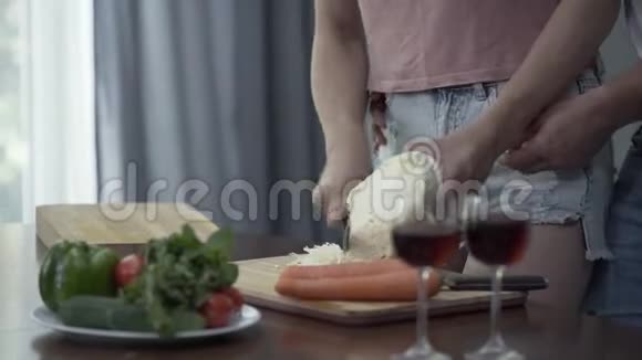 一个穿着牛仔裤短裤的年轻女人正在切卷心菜准备浪漫的晚餐她丈夫从后面拥抱她玻璃视频的预览图