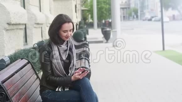 漂亮的女孩穿着黑色皮夹克和灰色围巾坐在长凳上看着电话他抬起头微笑着视频的预览图
