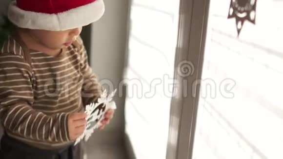 戴着圣诞帽的小宝宝在室内对着窗户玩圣诞装饰新年快乐圣诞快乐视频的预览图