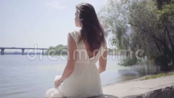 一位美丽迷人的少女一头深色长发穿着白色的夏装坐在海滨视频的预览图