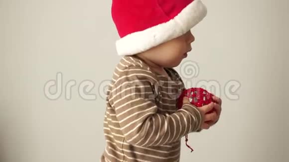 戴圣诞帽的可爱宝宝手里拿着一个红色的圣诞球准备圣诞晚会快乐的诺埃尔视频的预览图