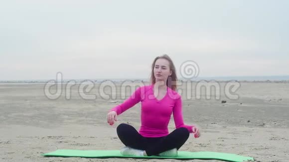 运动的年轻美丽的女孩在荒芜的海边沙滩上做体操坐在绿色的地毯上双手在不同的地方倾斜视频的预览图