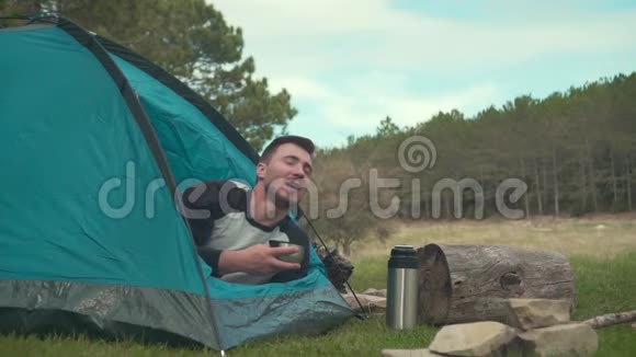一个年轻人躺在森林里的蓝色帐篷里喝着热水瓶里的茶享受着这一刻视频的预览图