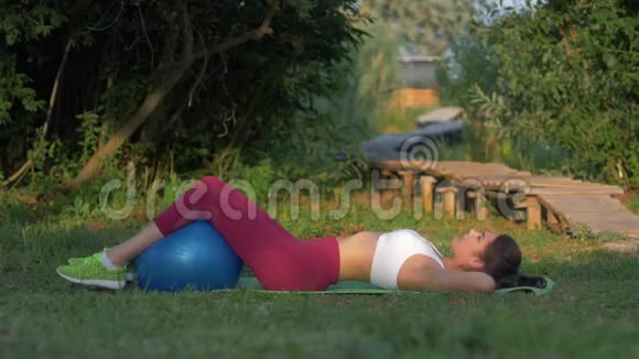年轻的健身女孩在自然的绿色草坪上用仰卧位的按压和双腿锁定大健身球视频的预览图