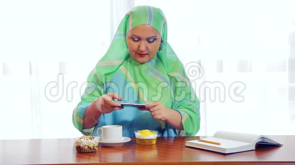 一个年轻的穆斯林妇女戴着绿色围巾在咖啡馆里拍照糕点在电话里视频的预览图