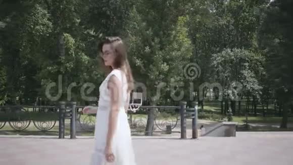 可爱的年轻女孩一头长长的黑发戴着太阳镜穿着一件白色的夏季时装长裙走在街上视频的预览图