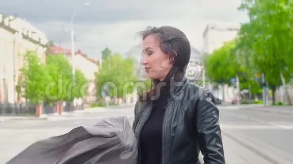 在一个阳光明媚的夏天穿着黑色皮夹克的苗条迷人女孩沿着城市的街道散步视频的预览图