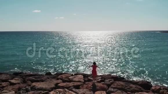 黑色的女性剪影穿着红色的衣服手臂分开在空中站在岩石海滩码头上对抗太阳太阳之路蓝色的倒影视频的预览图