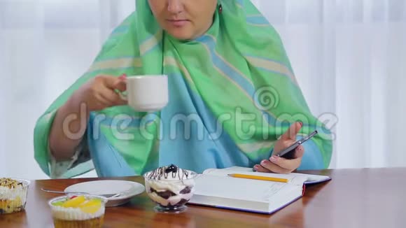 一个穿着浅绿色围巾的年轻穆斯林妇女在一家咖啡馆里用电话和日记远程工作视频的预览图