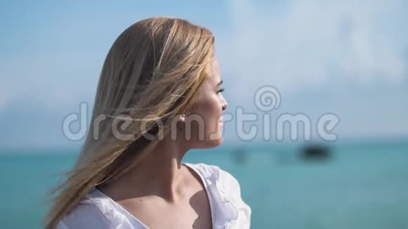 一位金发女郎穿着一件白色上衣站在海边或大海上凝视着大海的近景视频的预览图