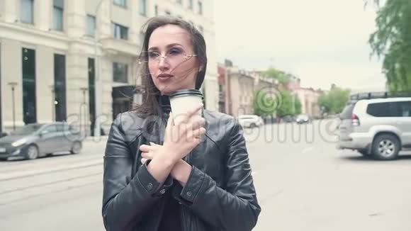 漂亮的年轻女孩穿着黑色皮夹克手里拿着咖啡整理她的头发戴太阳镜在背景通行证里视频的预览图