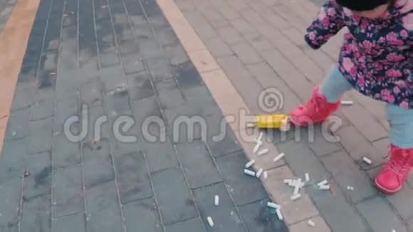 一个穿着五颜六色的粉红色夹克的可爱的小女孩散落在公园里儿童游乐场附近人行道上视频的预览图