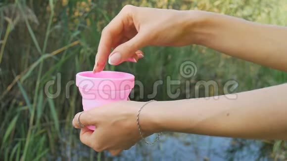 女性的手正在展示如何使用粉红色硅胶月经杯站在绿色自然背景与湖泊妇女视频的预览图