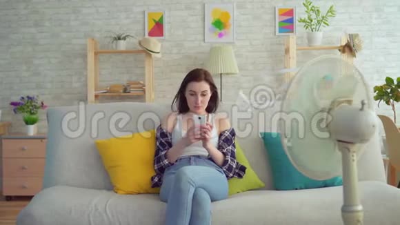 年轻女性使用智能手机坐在电风扇前的沙发上从热中逃脱视频的预览图