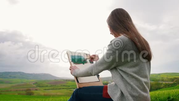 一个年轻的女人坐在绿色的田野中间画画头发随风飘动视频的预览图
