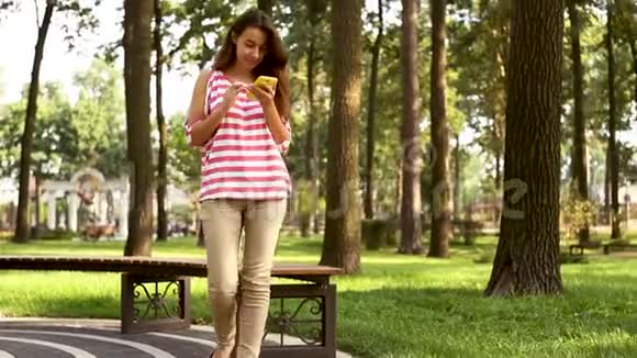 美丽的女孩在城市公园散步使用智能手机年轻的女人拿着设备和触摸屏幕夏天视频的预览图