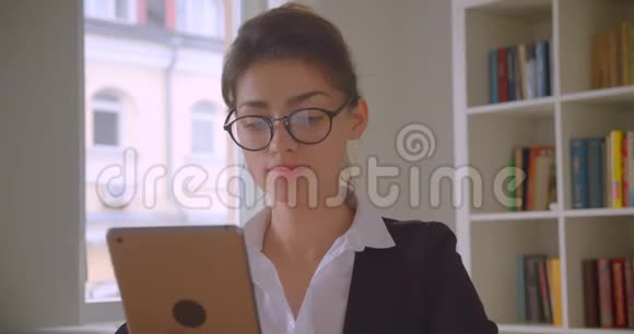 一位年轻漂亮的白种人女商人戴着眼镜在办公室室内的平板电脑上浏览特写镜头视频的预览图