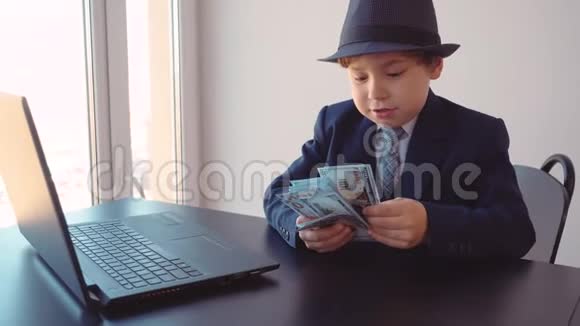 小男孩看起来像一个戴着帽子和西装的商人坐在他的办公室里在笔记本电脑附近数视频的预览图