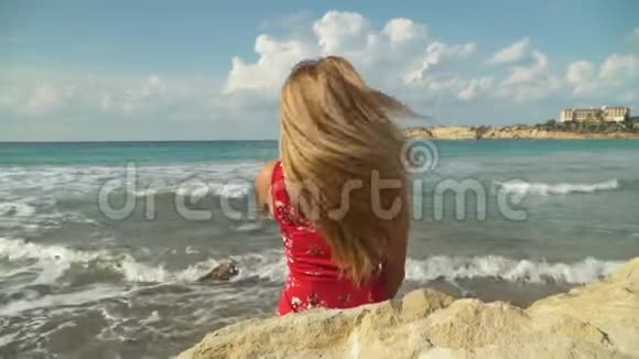 金发女人坐在岩石上看着挥手的大海可爱的女孩花时间在海边休闲视频的预览图