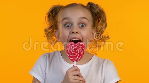 可爱的女孩用辫子舔心形糖孤立的黄色背景视频的预览图