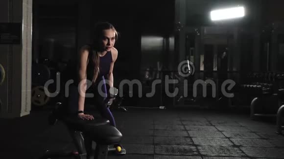 年轻女孩在健身房锻炼健身举起一只手哑铃靠在健身房的长凳上视频的预览图