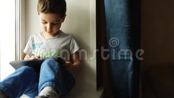 可爱的男孩玩平板电脑坐在房间的窗台上视频的预览图