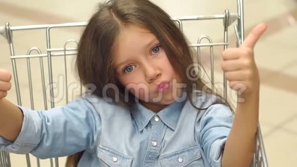 一个滑稽的小女孩坐在超市的购物车里伸出大拇指视频的预览图