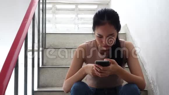 前面看到一个悲伤的女孩正在阅读网络欺凌的电话信息双手捧着脸坐在老公寓的楼梯上悲伤的女孩哭泣戏剧视频的预览图
